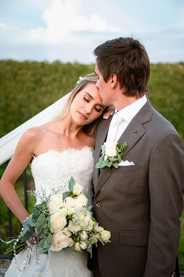 La novia y el novio están muy juntos, sonriendo para una foto de boda, vestidos con atuendos formales y con un fondo romántico.